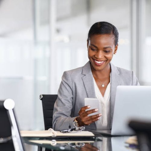 African american businesswoman using smartphone while working on laptop in office. Smiling mature black business woman checking phone while working. Successful woman entrepreneur browsing on phone in office.